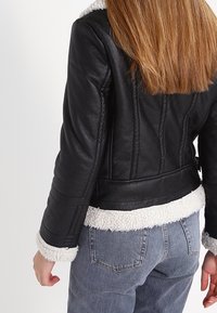 Svart läderjacka med vit konstpäls i kragen och foder, skräddarsydd passform, dragkedjestängning och ribbade ärmslut. Bärs med grå jeans.