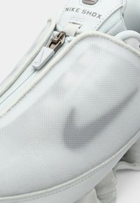 Παπούτσια Nike Shox με λευκή επένδυση από διχτυωτό υλικό, με κεντρικό φερμουάρ και γκρι λογότυπο Nike. Η σόλα διαθέτει ορατούς αποσβεστήρες κρούσης.