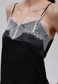 Svart sateng camisole med hvit blonderkant langs halsen, med tynne justerbare stropper og en glatt tekstur.
