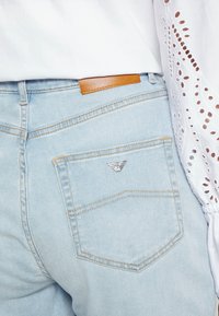 Ljust blå denimjeans med en läderetikett på midjan, med en silverlogotyp på bakfickan och synliga sömnadsdetaljer.