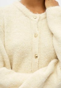 Strickcardigan in Creme aus weichem Material, mit tiefem V-Ausschnitt, fünf goldfarbenen Knöpfen und gerippten Bündchen für zusätzliche Textur.