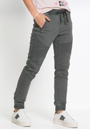 Persona che indossa pantaloni jogger testurizzati grigio scuro con vita con coulisse, tasche con zip e sneakers bianche con suola platform su sfondo bianco.