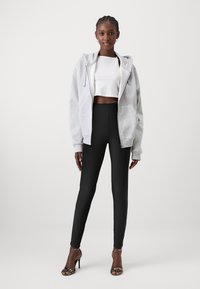 Un sweat à capuche gris zippé sur un crop top blanc, assorti à un legging noir taille haute et des talons noirs à lanières, se tenant sur un fond neutre.