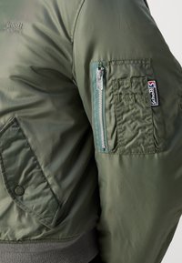 Veste bomber vert olive avec une finition lisse, une poche avant à pression et une poche zippée sur le bras avec étiquette de marque. Bords-côtes texturés.