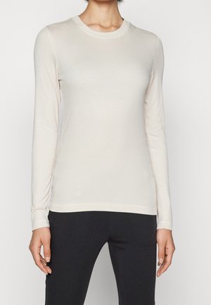 Kobieta ubrana w dopasowaną, kremową, prążkowaną bluzkę z długim rękawem oraz czarne spodnie, stojąca na jednolitym jasnym tle.