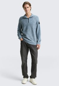 Blauwe gebreide pullover met een ritskraag, lange mouwen, textuurstof en geribbelde zoom. Gecombineerd met donkere jeans en witte sneakers.