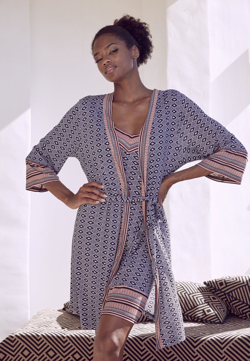 Blau und weiß gemustertes Wickelkleid mit einem Bindegürtel und V-Ausschnitt. Hat lange Ärmel mit kontrastierendem pinkem Saum. Getragen über einem passenden Oberteil.