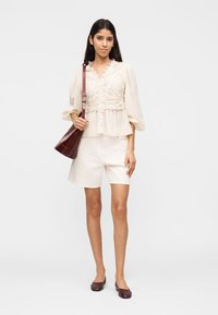 Γυναίκα που φοράει μπλούζα από εκρού δαντέλα και λευκά σορτς, κρατώντας σκούρα καφέ τσάντα ώμου και φορώντας καφέ μπαλαρίνες, στέκεται στο λευκό φόντο.