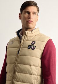 Chaleco acolchado beige con cremallera frontal, que presenta un cuello azul marino y tres parches en forma de escudo. Lleva puesto sobre una camiseta de manga larga roja. Textura suave.