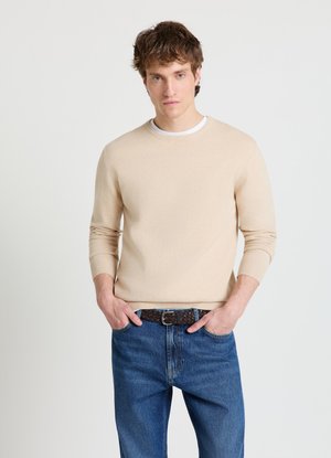 Giovane uomo con maglione beige girocollo, jeans blu e cintura intrecciata scura, posa con le mani in tasca su sfondo chiaro e uniforme.