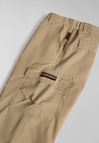 Beige cargo bukser med en glat tekstur, der har flere lommer og et mærke på bagsiden. Elastisk talje inkluderet.