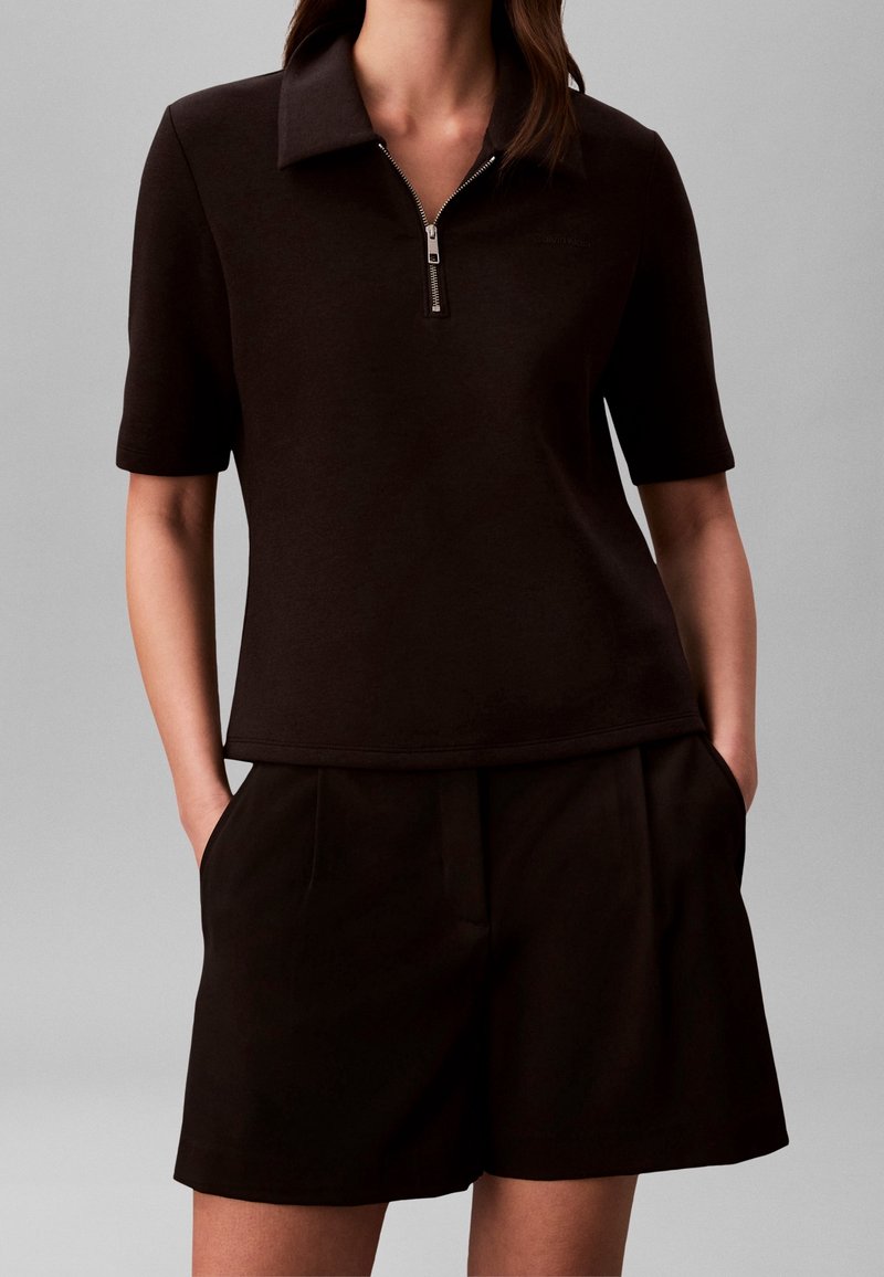 Femme portant un polo noir à manches courtes zippé et un short noir ajusté, les mains dans les poches, sur un fond uni.