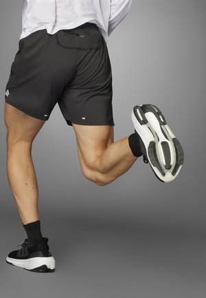 Persoon in zwarte sportbroek en zwarte hardloopschoenen, vastgelegd in het midden van een spron tegen een grijze achtergrond.