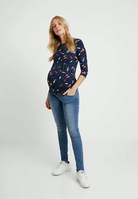 Mulher grávida com cabelo loiro longo está de pé com uma mão no bolso, vestindo uma blusa floral azul marinho, jeans azuis e ténis brancos, em um fundo plain.