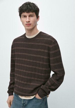 Giovane uomo con maglione a righe marrone scuro sopra una camicia beige e jeans blu, in piedi contro uno sfondo chiaro e uniforme.