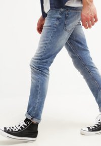 Light blue denim jeans with a slim fit, featuring faded sections, a subtle whisker pattern, and rolled cuffs, paired with black high-top sneakers.