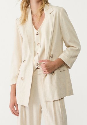 Vrouw die een crèmekleurige dubbelgeknoopte blazer draagt, een bijpassend gilet met knopen en losse broek, terwijl ze met één hand de blazer rechtzet.