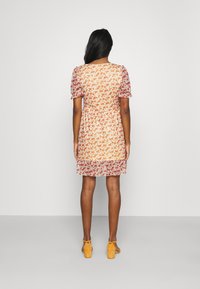 Blumenkleid mit einem gelb-roten Muster, kurzen Puffärmeln, tailliertem Bund und Rüschensaum. Getragen mit gelben Absatzsandalen.