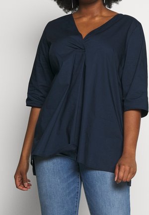 Mujer con una blusa holgada de color azul marino oscuro con cuello en V y mangas tres cuartos, combinada con vaqueros azul claro, frente a un fondo liso.