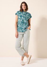 Camicetta azzurra a maniche corte con bottoni e motivo decorativo, abbinata a jeans larghi blu chiaro e sneakers bianche.