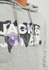 Sudadera gris con gran texto blanco "JACK & JONES" y un gráfico de globo púrpura, con un patrón de rejilla en los continentes delineados en negro.