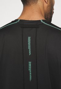 Černé sportovní tričko s zeleným prošitím podél švů a potiskem na zádech, navržené pro pohodlí a odvod vlhkosti.
