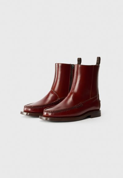 Bottines en cuir rouge avec une texture lisse, fermeture éclair sur le côté et tirette contrastante en brun. Présente un motif de piqûre décoratif.