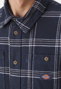 Camicia a quadri blu navy realizzata in tessuto morbido, con colletto abbottonato, tasche sul petto e una patch con logo dai contrasti crema e sabbia.