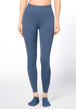 Woman wearing high-waisted, seamless blue leggings and matching footed socks, standing against a plain white background.