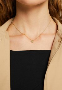 Collana in oro con un piccolo pendente a forma di fiore con accenti verdi, indossata sopra una maglietta nera e un blazer beige.
