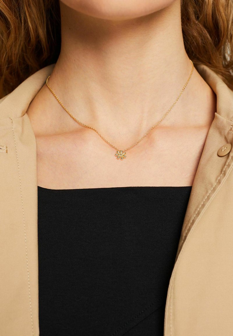 Collana in oro con un piccolo pendente a forma di fiore con accenti verdi, indossata sopra una maglietta nera e un blazer beige.