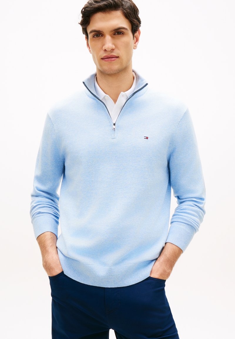 Jeune homme portant un pull à demi-fermeture éclair bleu clair par-dessus une chemise blanche à col, pantalon bleu foncé, debout les mains dans les poches, fond neutre.