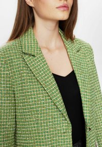 Grön tweedblazer med ett strukturerat rutor-mönster, med notched krage och enkelknappstängning, bärs över en svart topp.