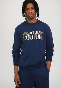 Mornarsko modra dukserica iz mehkega blaga z drznim zlatim in belim logotipom "VERSACE JEANS COUTURE" na prsi. Riblji manšeti in rob.