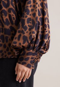 Blusa a stampa leopardata marrone e nera con maniche a sbuffo e polsini arricciati, dotata di bottoni lungo i bordi dei polsini. Tessuto dalla texture liscia.