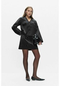 Sorte satin kjole med V-udskæring, lange ærmer og blomsterlignende udsmykninger. Båret med sorte flade sko og holder en sort taske.