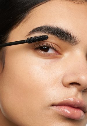 Scovolino del mascara nero che applica il prodotto su sopracciglia spesse e scure. La pelle mostra una texture liscia con una finitura naturale e leggermente lucida.