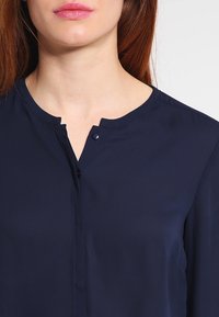 Mørkeblå bluse lavet af glat stof, med mandarin-krave, frontknaplukning og en beskeden halsudskæring.