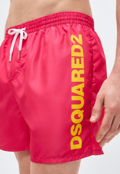 Κοντινό πλάνο κόκκινων μαγιό με λευκό κορδόνι και κίτρινο κάθετο λογότυπο "DSQUARED2" στο πλευρό, που φοράει ένα άτομο.