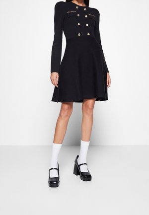 Femme portant une robe noire à manches longues avec des boutons dorés, des chaussettes blanches et des chaussures Mary Jane noires à talons blocs.