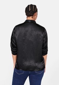 Camicia nera di raso con maniche lunghe e colletto, caratterizzata da una texture leggermente sgualcita e un taglio dritto nella parte inferiore.