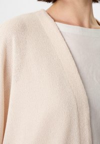 Světle růžový kardigan s žebrovanou strukturou, designem s otevřeným předním dílem a lehkým materiálem. Převlečený přes béžový top s jednoduchým výstřihem.