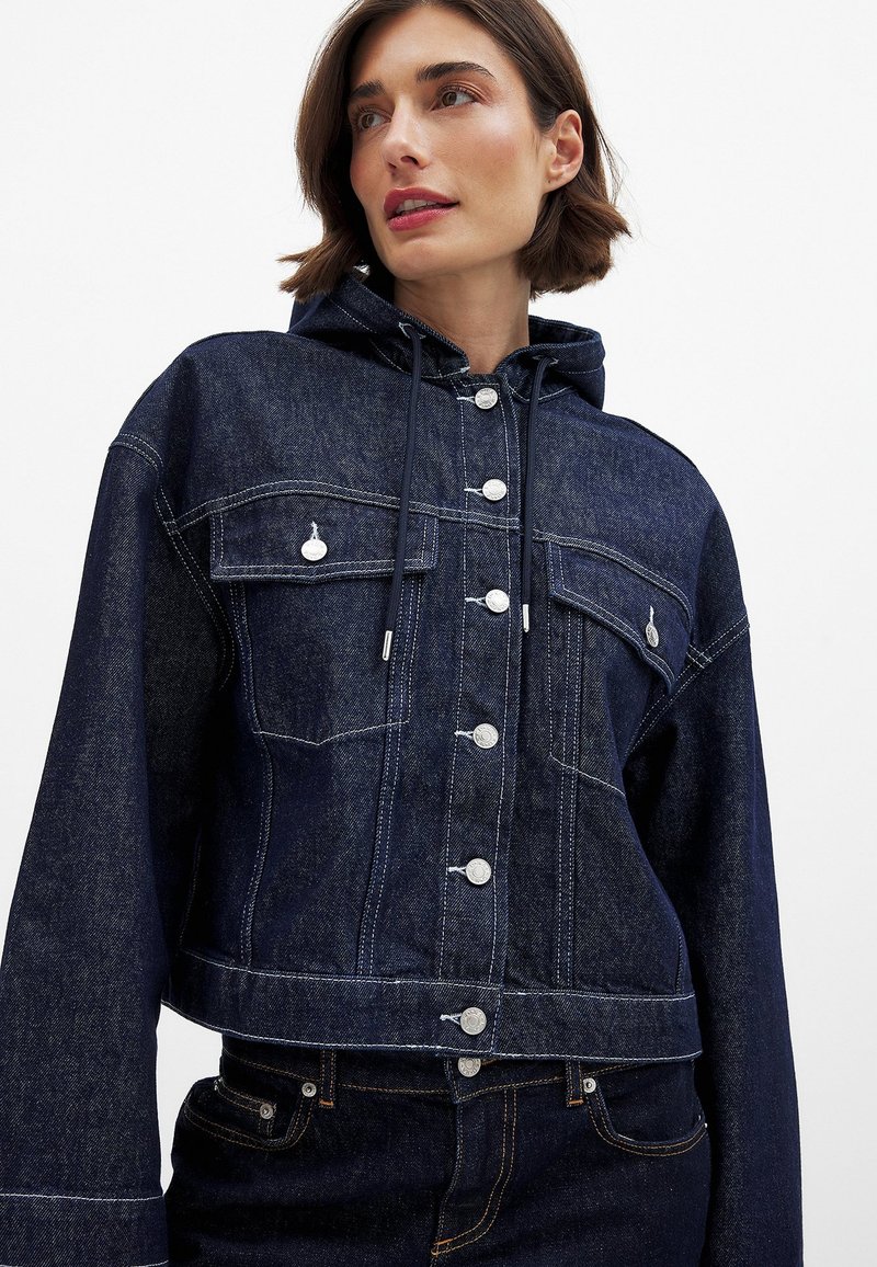 Izgriezta tumši zila džinsu jaka ar kapuci, sudraba pogām un divām krūšu kabatām. Iezīmējas ar kontrastējošu balto šuvumu un aukliņu ar slēgšanu.