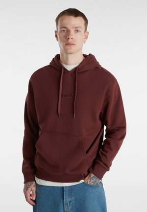 Bordeauxrode hoodie van een zachte stof met een voorkruiszak, een capuchon met trekkoord en een geborduurd logo. Draagt over een wit shirt; gecombineerd met blauwe jeans.