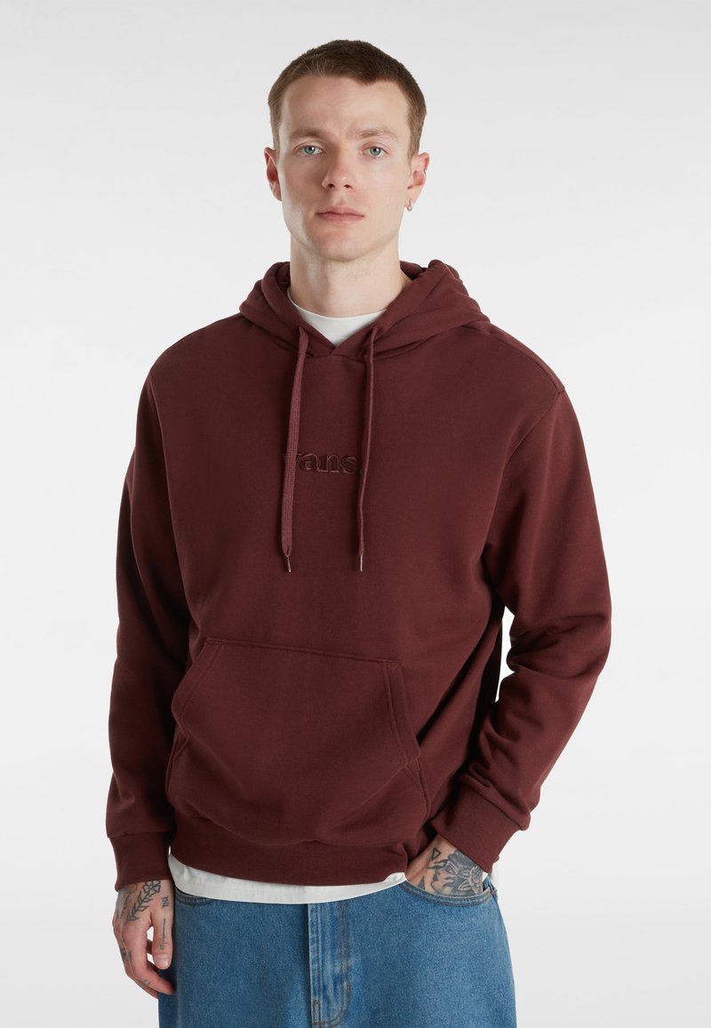 Felpa maroon in un tessuto morbido con tasca frontale, cappuccio con cordino e logo ricamato. Indossata sopra una maglietta bianca; abbinata a jeans blu.