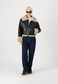 Veste cropped en cuir noir avec col et poignets en shearling beige, dotée d'une fermeture éclair à l'avant et de deux poches latérales, portée avec un jean noir à jambes larges.