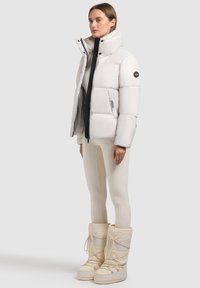 Giacca bianca gonfia con colletto alto e chiusura con zip, abbinata a leggings beige e stivali leggeri. La giacca presenta un'etichetta con logo e un design elegante.