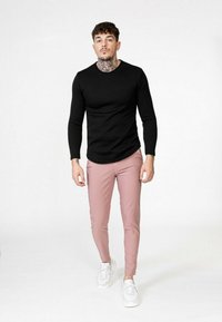 Haut ajusté à manches longues noir côtelé associé à un pantalon slim rose clair et des baskets blanches. Fond uni mettant en valeur la tenue.