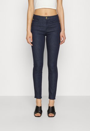 Kobieta ubrana w ciemnoniebieskie wąskie dżinsy, biały krótki top i czarne otwarte sandały na obcasie, stoi na jednolitym tle.