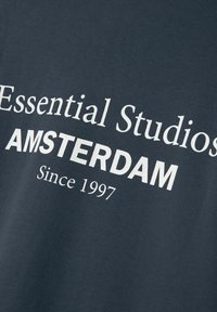 Grå bomullströja med vit text: "Essential Studios AMSTERDAM Sedan 1997." Enkel rund halsringning med mjuk textur.
